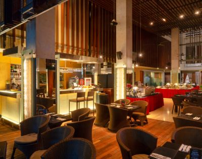 Spacious, warmly lit restaurant with modern decor, featuring cushioned wicker chairs and elegantly set tables. A buffet area in the background adds a cozy ambiance.