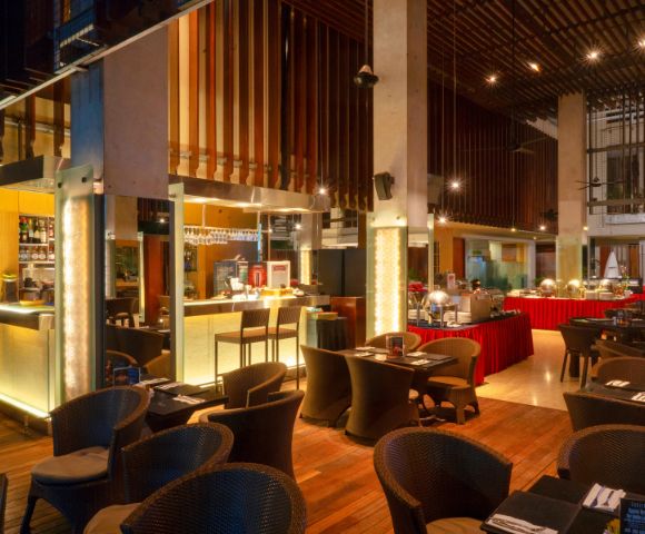 Spacious, warmly lit restaurant with modern decor, featuring cushioned wicker chairs and elegantly set tables. A buffet area in the background adds a cozy ambiance.