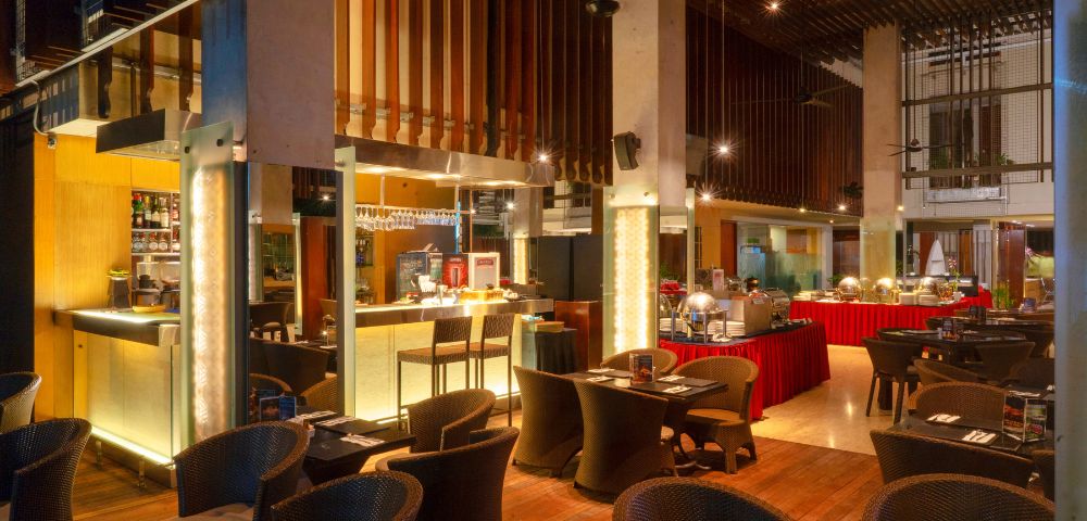 Spacious, warmly lit restaurant with modern decor, featuring cushioned wicker chairs and elegantly set tables. A buffet area in the background adds a cozy ambiance.