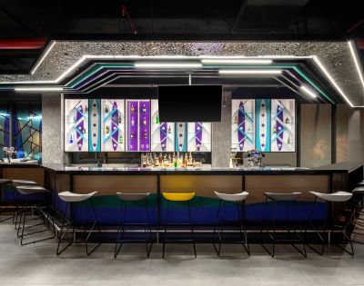 Modern bar interior with geometric patterns and vivid colors on backlit panels. Sleek stools line the counter, creating a vibrant, urban atmosphere.