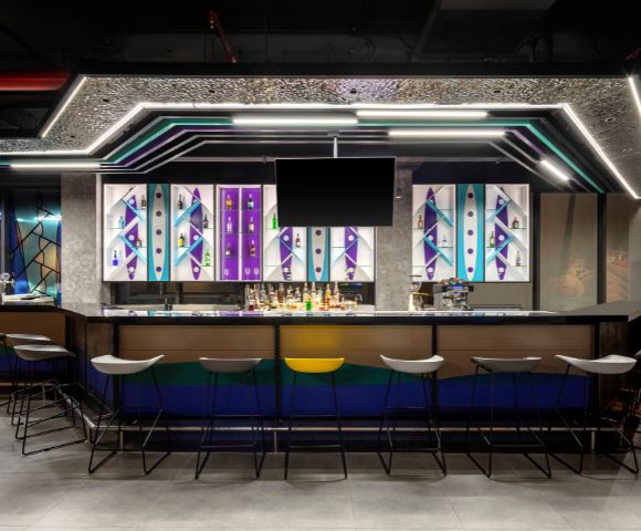 Modern bar interior with geometric patterns and vivid colors on backlit panels. Sleek stools line the counter, creating a vibrant, urban atmosphere.