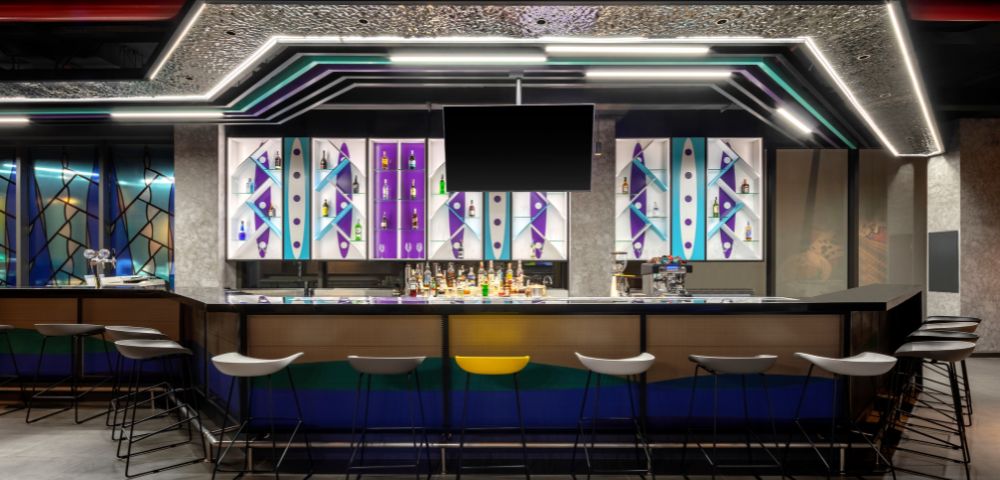 Modern bar interior with geometric patterns and vivid colors on backlit panels. Sleek stools line the counter, creating a vibrant, urban atmosphere.
