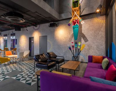 Modern lounge with colorful furniture including purple and yellow couches, geometric flooring, and kayak decor; bright and lively atmosphere.