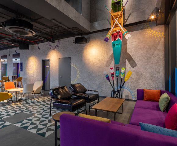 Modern lounge with colorful furniture including purple and yellow couches, geometric flooring, and kayak decor; bright and lively atmosphere.