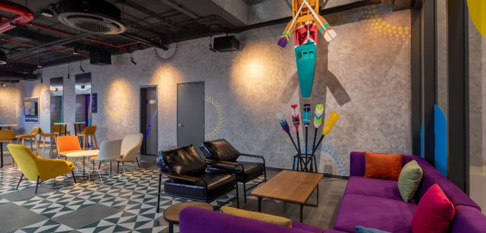 Modern lounge with colorful furniture including purple and yellow couches, geometric flooring, and kayak decor; bright and lively atmosphere.