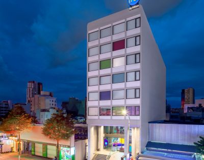 Modern hotel building at dusk, with a colorful window pattern and vibrant street below. The sky is dark, setting a calm urban atmosphere.