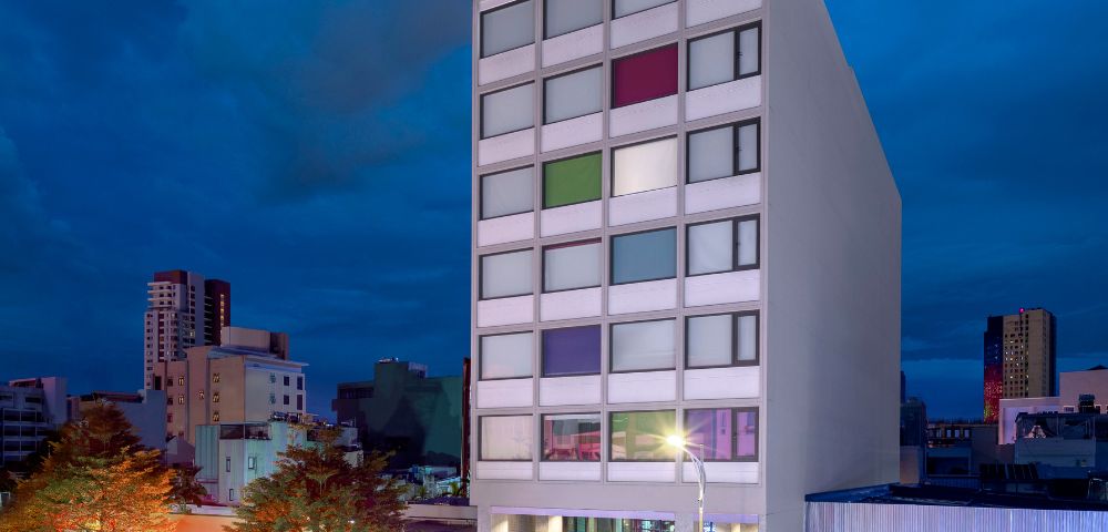 Modern hotel building at dusk, with a colorful window pattern and vibrant street below. The sky is dark, setting a calm urban atmosphere.