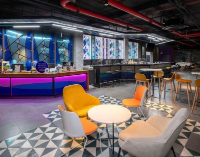 Stylish cafeteria with modern decor features colorful chairs around round tables, vibrant blue and purple counters, geometric floor pattern, and warm lighting.