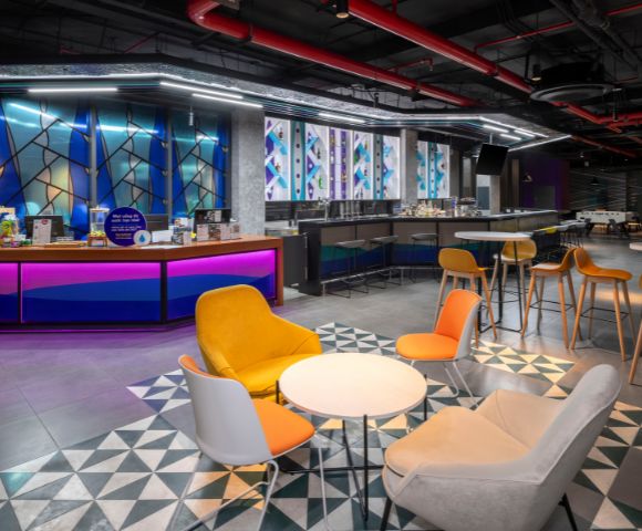 Stylish cafeteria with modern decor features colorful chairs around round tables, vibrant blue and purple counters, geometric floor pattern, and warm lighting.