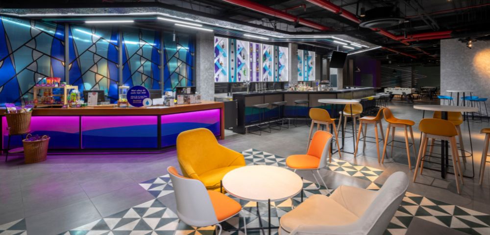 Stylish cafeteria with modern decor features colorful chairs around round tables, vibrant blue and purple counters, geometric floor pattern, and warm lighting.