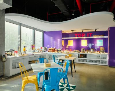 Bright, modern breakfast area with purple walls and white countertops. Blue and yellow chairs surround tables. Natural light streams in, creating a welcoming atmosphere.
