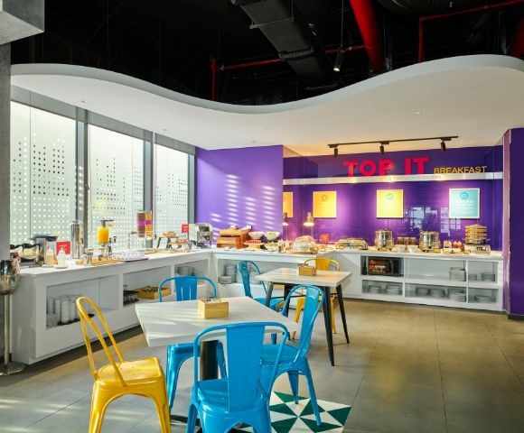 Bright, modern breakfast area with purple walls and white countertops. Blue and yellow chairs surround tables. Natural light streams in, creating a welcoming atmosphere.