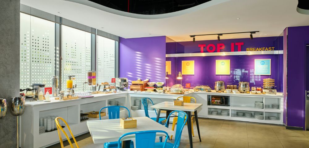 Bright, modern breakfast area with purple walls and white countertops. Blue and yellow chairs surround tables. Natural light streams in, creating a welcoming atmosphere.