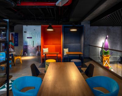 A modern, vibrant workspace featuring colorful alcoves with tables, blue and orange chairs, dark ceiling, and playful decor, conveying creativity and energy.