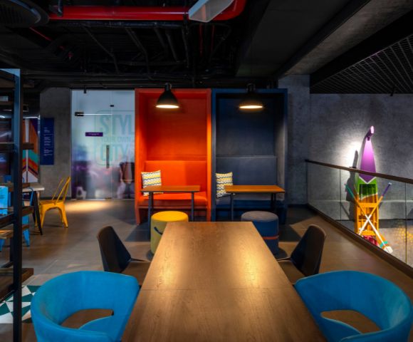 A modern, vibrant workspace featuring colorful alcoves with tables, blue and orange chairs, dark ceiling, and playful decor, conveying creativity and energy.