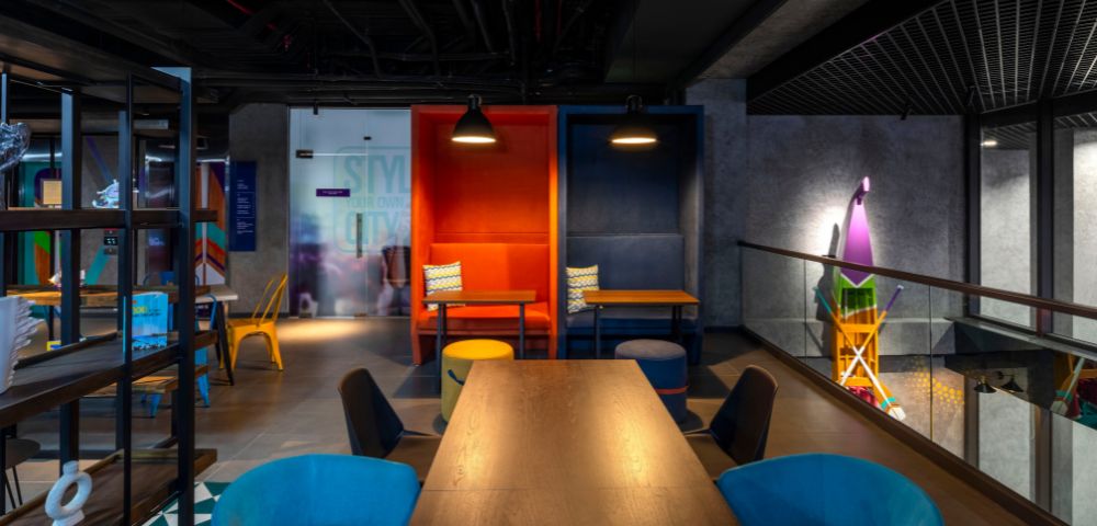 A modern, vibrant workspace featuring colorful alcoves with tables, blue and orange chairs, dark ceiling, and playful decor, conveying creativity and energy.