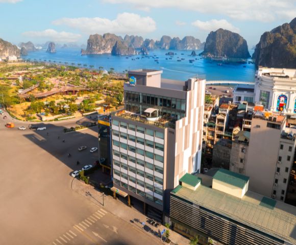 Aerial view of a coastal city with a prominent multi-story building in the foreground, lush green park, and scenic limestone islands under a bright sky.