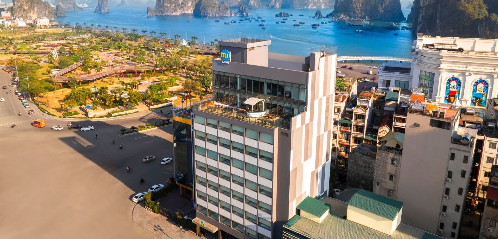 Aerial view of a coastal city with a prominent multi-story building in the foreground, lush green park, and scenic limestone islands under a bright sky.