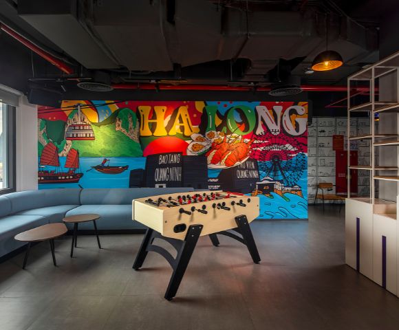 Modern lounge with a foosball table and colorful mural of Ha Long Bay. A curved sofa and round tables add a relaxed, playful atmosphere.