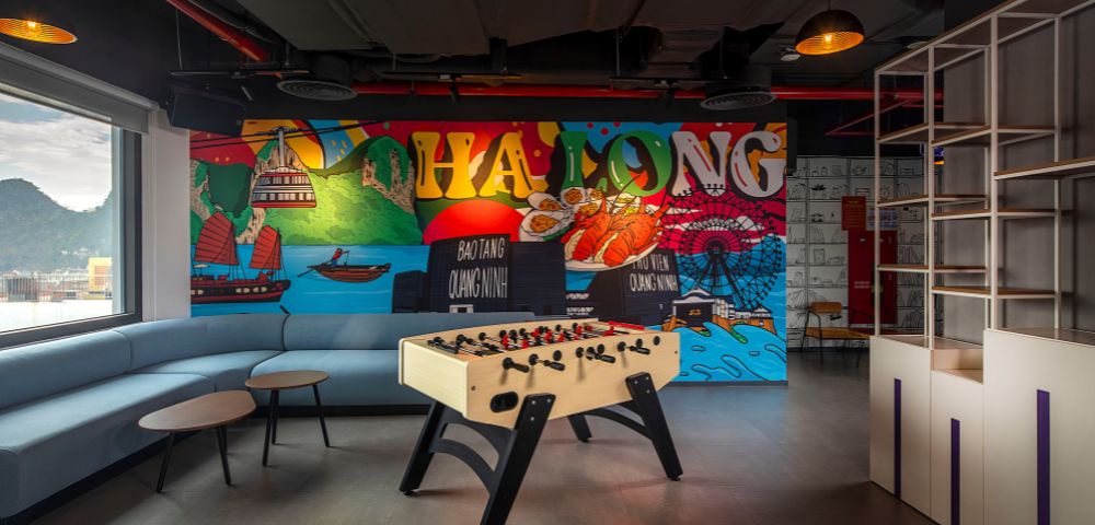 Modern lounge with a foosball table and colorful mural of Ha Long Bay. A curved sofa and round tables add a relaxed, playful atmosphere.