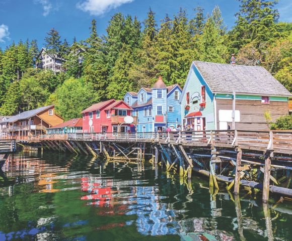 Colorful buildings on a wooden boardwalk line a serene waterfront, with lush green trees in the background, reflecting a peaceful, quaint atmosphere.