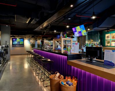 A modern bar with a long counter lit by purple LED lights, high black stools, digital menu screens, and snack baskets, creating a lively atmosphere.