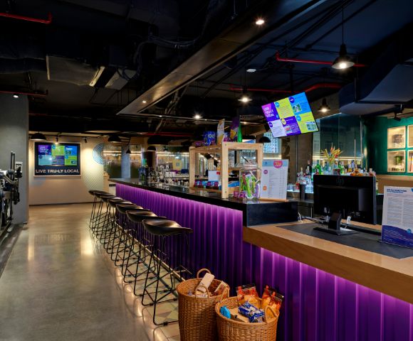A modern bar with a long counter lit by purple LED lights, high black stools, digital menu screens, and snack baskets, creating a lively atmosphere.