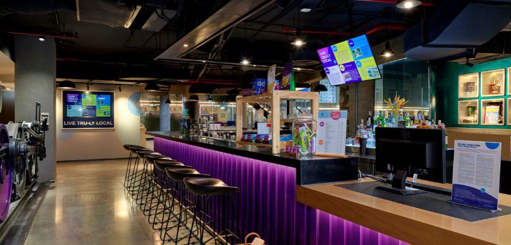 A modern bar with a long counter lit by purple LED lights, high black stools, digital menu screens, and snack baskets, creating a lively atmosphere.