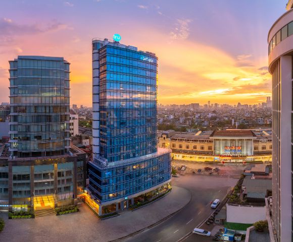A vibrant cityscape at sunset with glass buildings reflecting orange and pink hues. In the foreground, a curved road with cars. The scene is tranquil and modern.