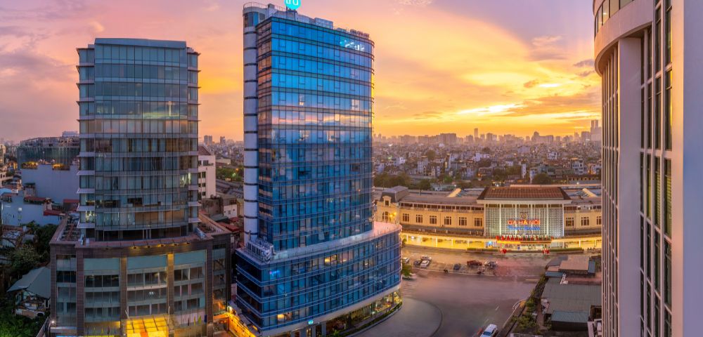 A vibrant cityscape at sunset with glass buildings reflecting orange and pink hues. In the foreground, a curved road with cars. The scene is tranquil and modern.