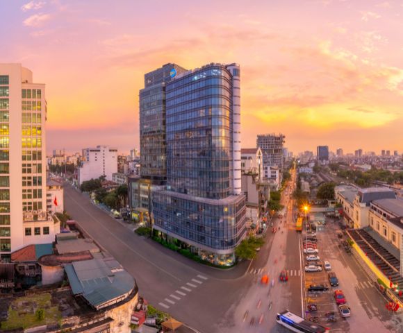 Aerial view of a cityscape at sunset, featuring a modern glass high-rise in the center. Streets and vehicles surround the glowing urban landscape.