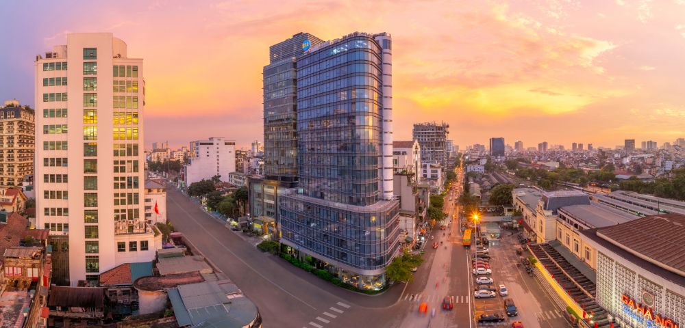 Aerial view of a cityscape at sunset, featuring a modern glass high-rise in the center. Streets and vehicles surround the glowing urban landscape.