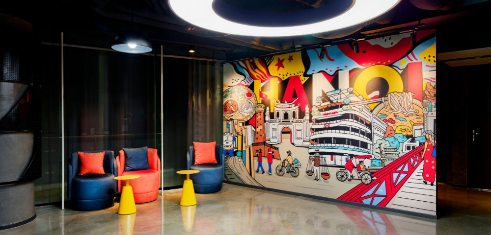 A modern room with bright, artistic Hanoi-themed graffiti on the wall. Two colorful chairs and small tables sit beneath a circular ceiling light.