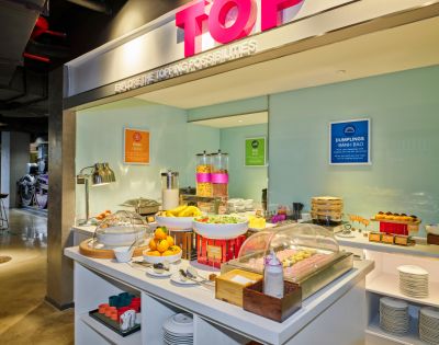 Spacious buffet area with a variety of breakfast items, including fruits, cereals, and drinks. Bright lighting creates a welcoming atmosphere.