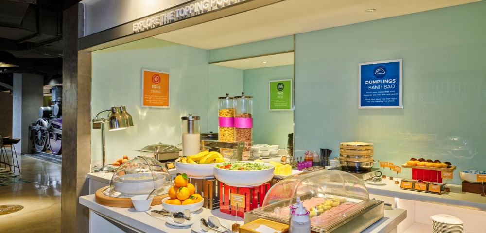 Spacious buffet area with a variety of breakfast items, including fruits, cereals, and drinks. Bright lighting creates a welcoming atmosphere.