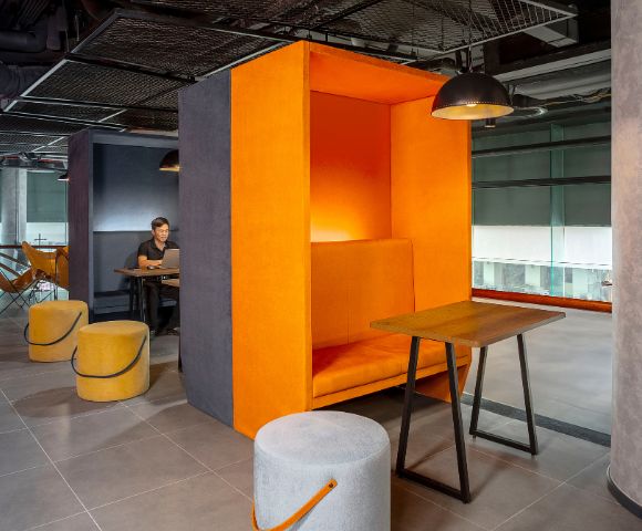 Modern office space with vibrant orange acoustic booth and seating, gray floors, pendant lighting, and a person working at a nearby table. Cozy and stylish.