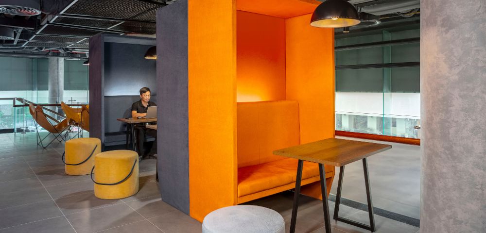 Modern office space with vibrant orange acoustic booth and seating, gray floors, pendant lighting, and a person working at a nearby table. Cozy and stylish.
