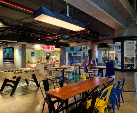 Modern office break area with vibrant decor, featuring a foosball table, colorful chairs, and a food counter. The setting feels lively and casual.