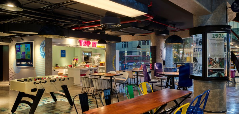 Modern office break area with vibrant decor, featuring a foosball table, colorful chairs, and a food counter. The setting feels lively and casual.