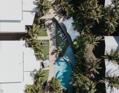 Aerial view of a tropical resort featuring a curved pool surrounded by palm trees and white-roofed buildings, conveying a relaxing and luxurious vibe.