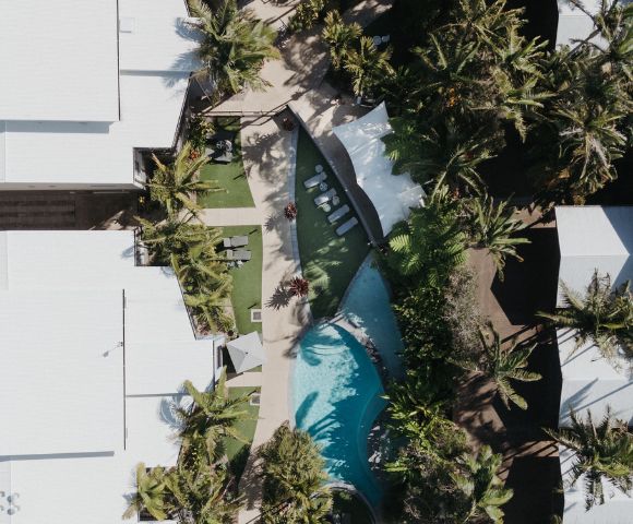 Aerial view of a tropical resort featuring a curved pool surrounded by palm trees and white-roofed buildings, conveying a relaxing and luxurious vibe.
