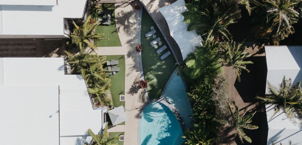 Aerial view of a tropical resort featuring a curved pool surrounded by palm trees and white-roofed buildings, conveying a relaxing and luxurious vibe.