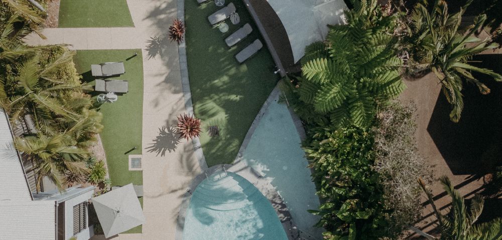 Aerial view of a serene outdoor pool area with curved edges, surrounded by lush green palm trees. Lounge chairs and sunshades add a relaxing vibe.