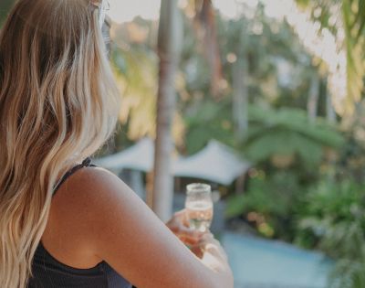 A woman with long blonde hair, facing away, holds a glass of champagne on a balcony overlooking lush greenery and a pool, conveying relaxation and tranquility.