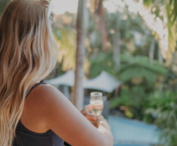 A woman with long blonde hair, facing away, holds a glass of champagne on a balcony overlooking lush greenery and a pool, conveying relaxation and tranquility.