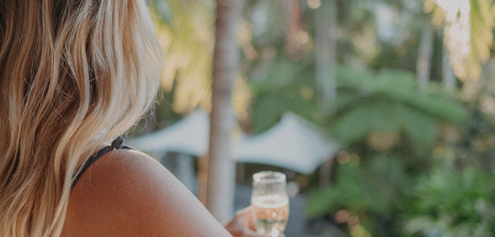 A woman with long blonde hair, facing away, holds a glass of champagne on a balcony overlooking lush greenery and a pool, conveying relaxation and tranquility.