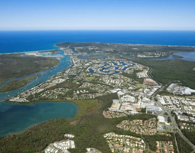 Aerial view of a coastal city with winding waterways, residential areas, and lush greenery. The ocean in the distance meets a clear blue sky.
