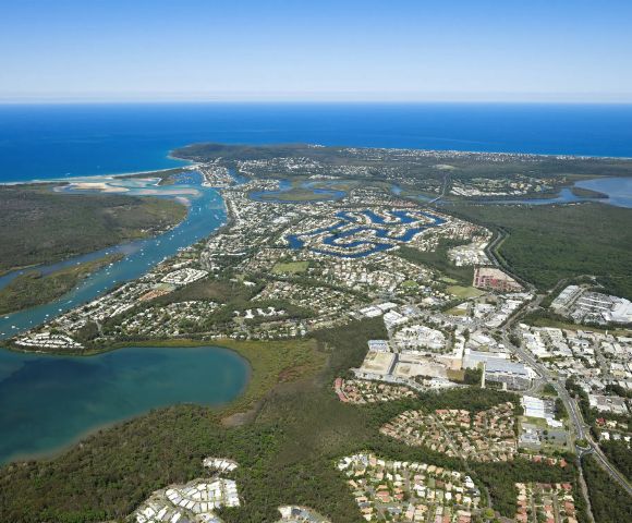 Aerial view of a coastal city with winding waterways, residential areas, and lush greenery. The ocean in the distance meets a clear blue sky.