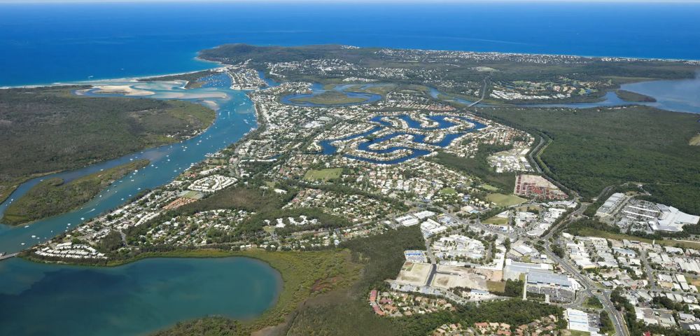 Aerial view of a coastal city with winding waterways, residential areas, and lush greenery. The ocean in the distance meets a clear blue sky.