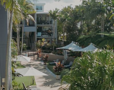 Lush tropical courtyard with palm trees and sunlit pathways. Lounge chairs and umbrellas create a serene, resort-like atmosphere beside modern buildings.